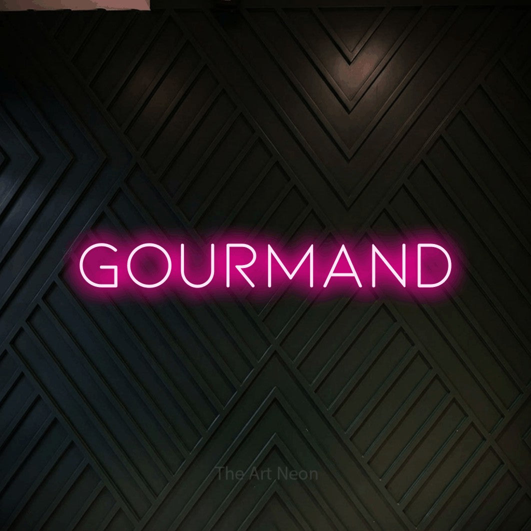 “Gourmand” LED neon sign in hot pink, with thin, widely spaced lettering, shown against a black chevron-patterned wood wall.