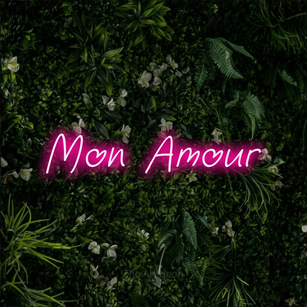 “Mon amour” LED neon sign in hot pink, with slanted handwritten lettering, shown against a dense green plant wall.