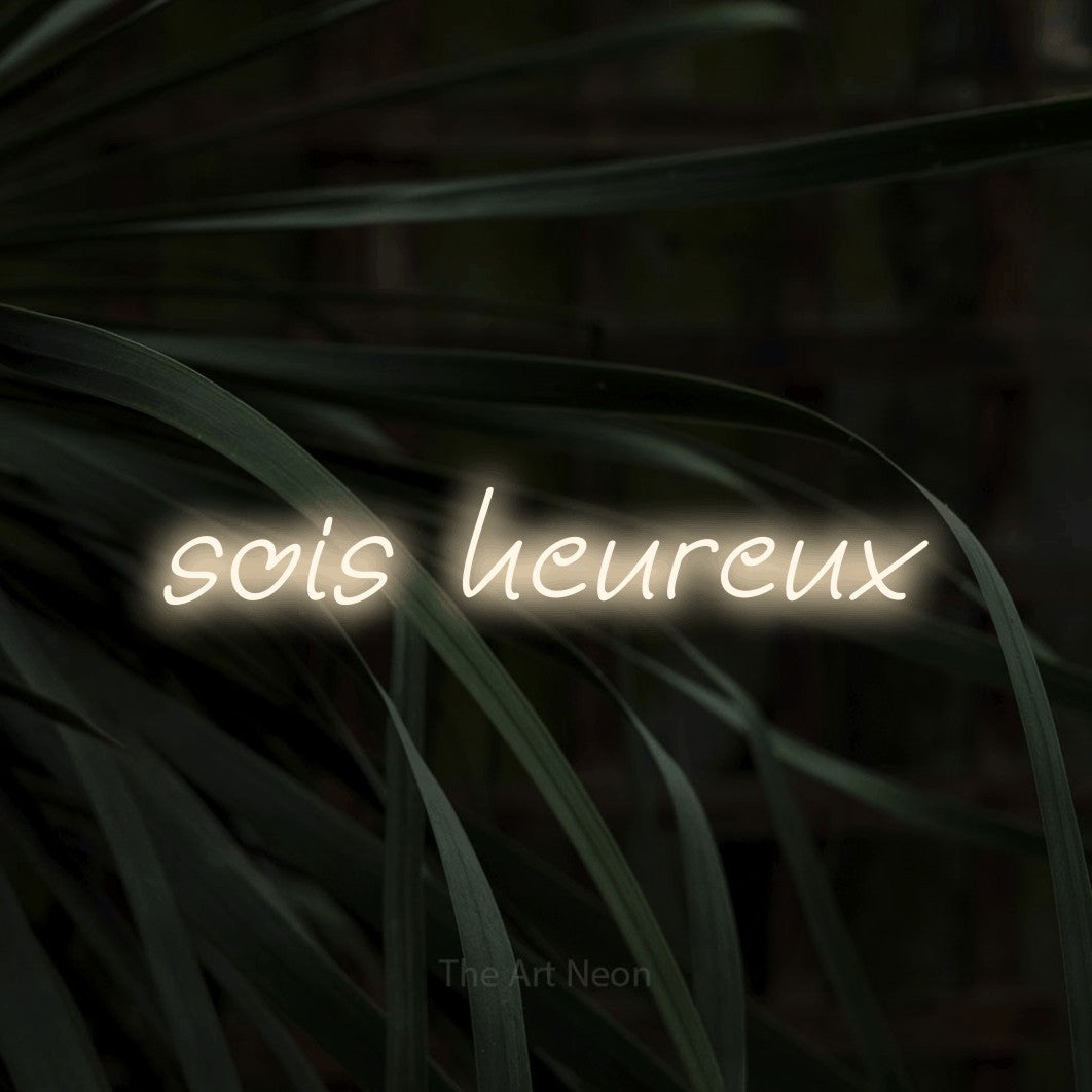 “Sois heureux” LED neon sign in warm white, with slanted handwritten lettering, shown against a close-up of dark foliage.
