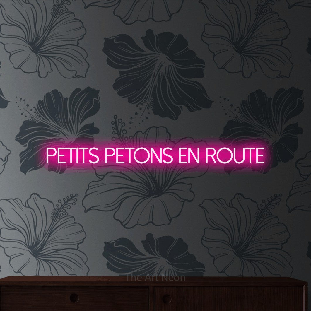 “Petits petons en route” LED neon sign in hot pink, with thin sans-serif lettering, shown against a grey floral-patterned wall.
