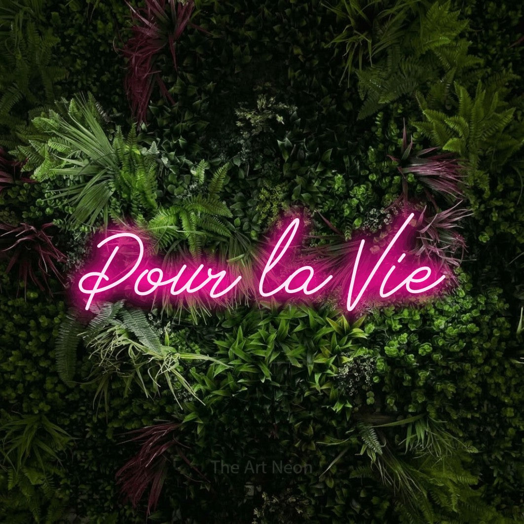 “Pour la vie” LED neon sign in hot pink, with cursive lettering, shown against a plant wall with pink flowers.