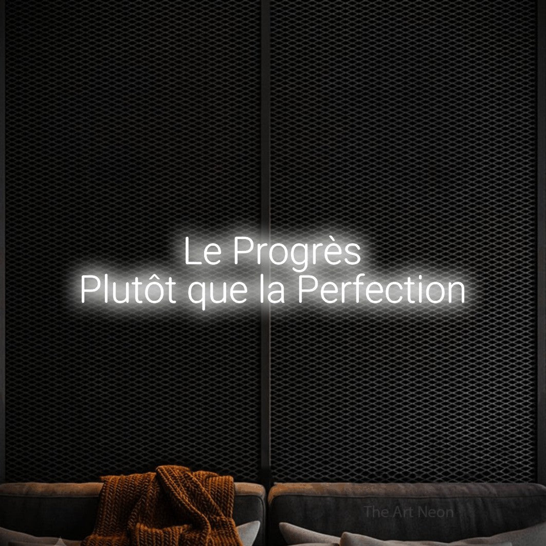 “Le progrès plutôt que la perfection” LED neon sign in cool white, with rounded sans-serif lettering across 2 lines, shown against a modern dark-toned seating area.
