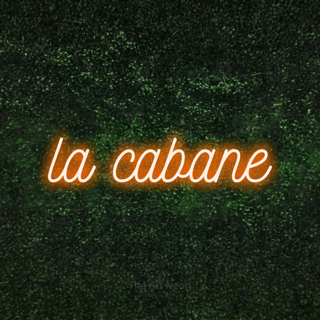 “La cabane” LED neon sign in orange, with ornate cursive lettering, shown against a textured green hedge.