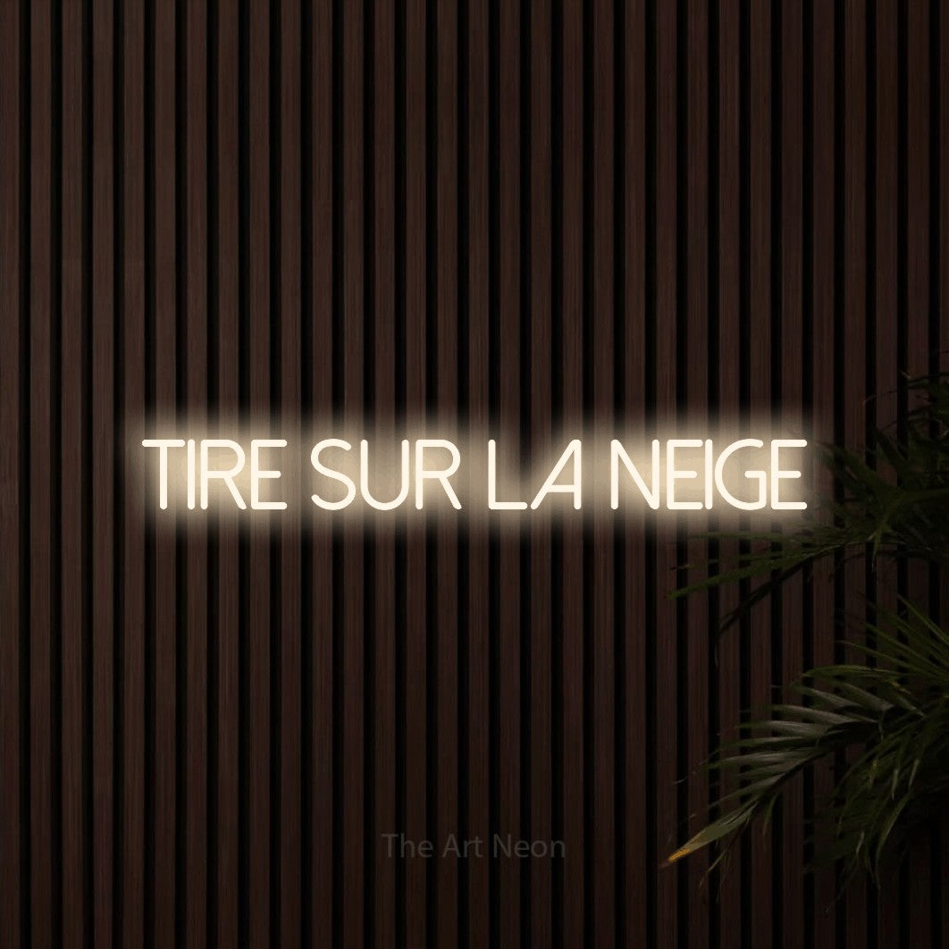 “Tire sur la neige” LED neon sign in warm white, with thin sans-serif lettering, shown against dark wooden wall slats.
