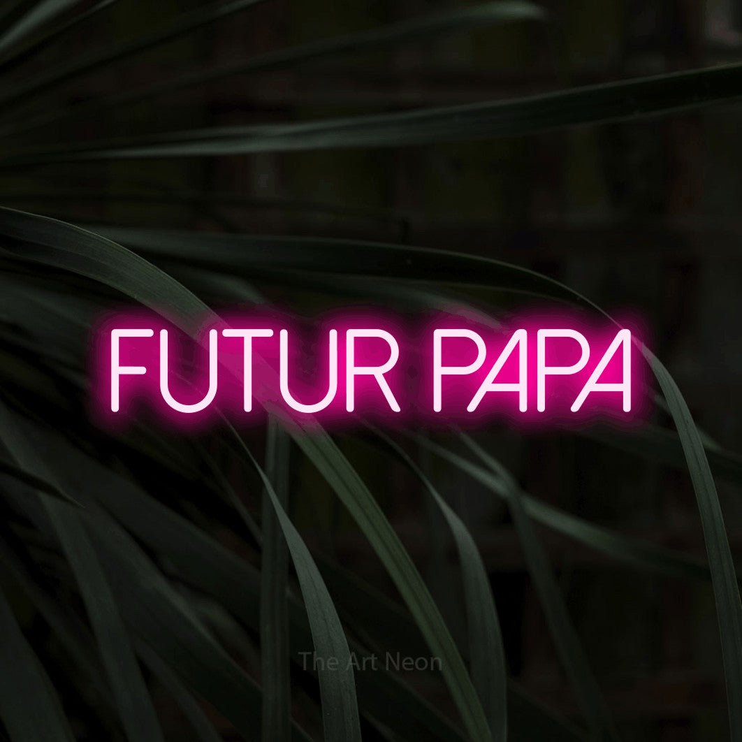 “Futur papa” LED neon sign in hot pink, with thin sans-serif lettering, shown against a close-up of dark foliage.
