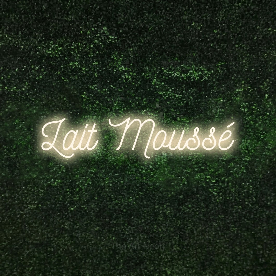 “Lait moussé” LED neon sign in warm white, with ornate cursive lettering, shown against a textured green hedge.