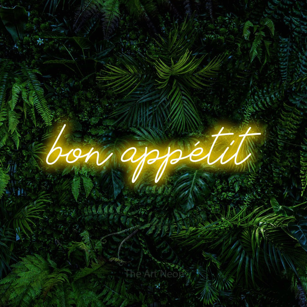 “Bon appétit” LED neon sign in yellow, with cursive lettering, shown against dense tropical foliage.