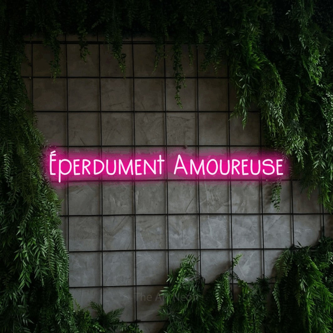 “Éperdument amoureuse” LED neon sign in hot pink, with narrow hand-drawn lettering, shown against an ivy-covered concrete grid.