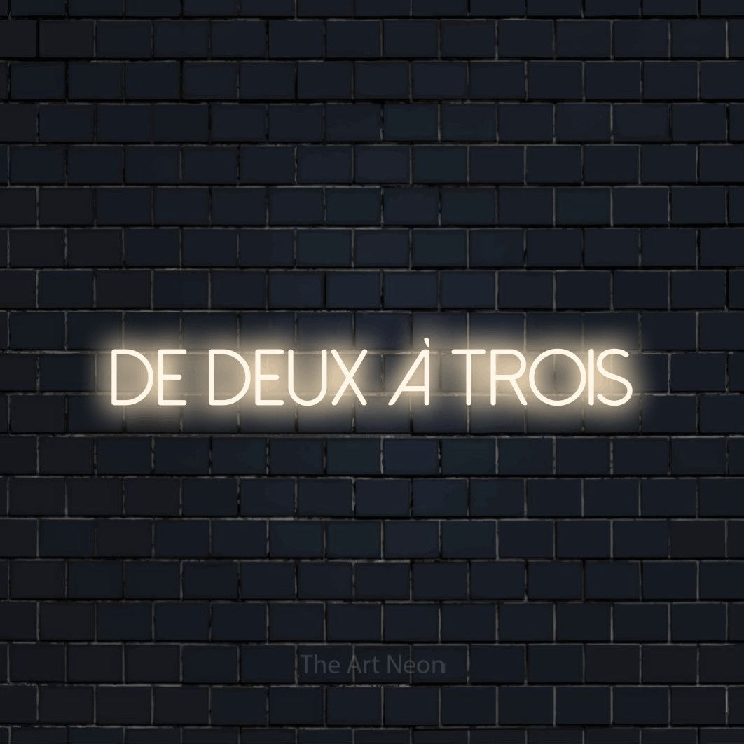 “De deux à trois” LED neon sign in warm white, with thin sans-serif lettering, shown against a dark navy brick wall.