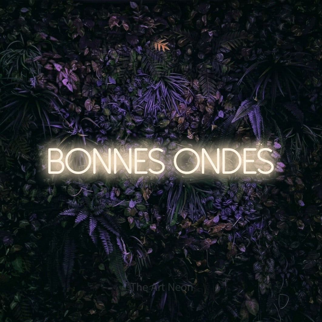 “Bonnes ondes” LED neon sign in warm white, with thin sans-serif lettering, shown against a dark wall with purple flowers.