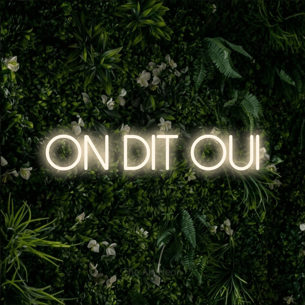 “On dit oui” LED neon sign in warm white, with thin sans-serif lettering, shown against a dense green plant wall.