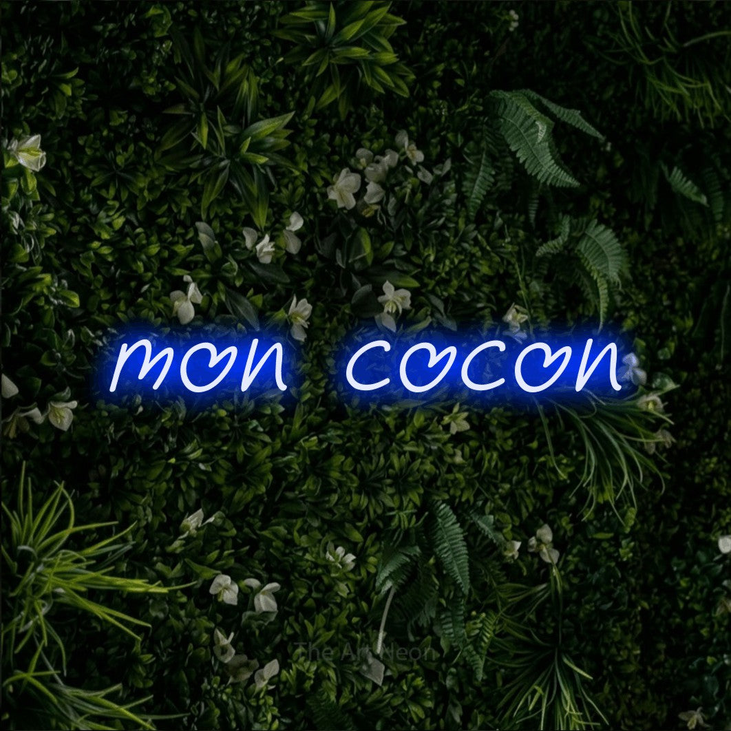 “Mon cocon” LED neon sign in blue, with slanted handwritten lettering, shown against a dense green plant wall.