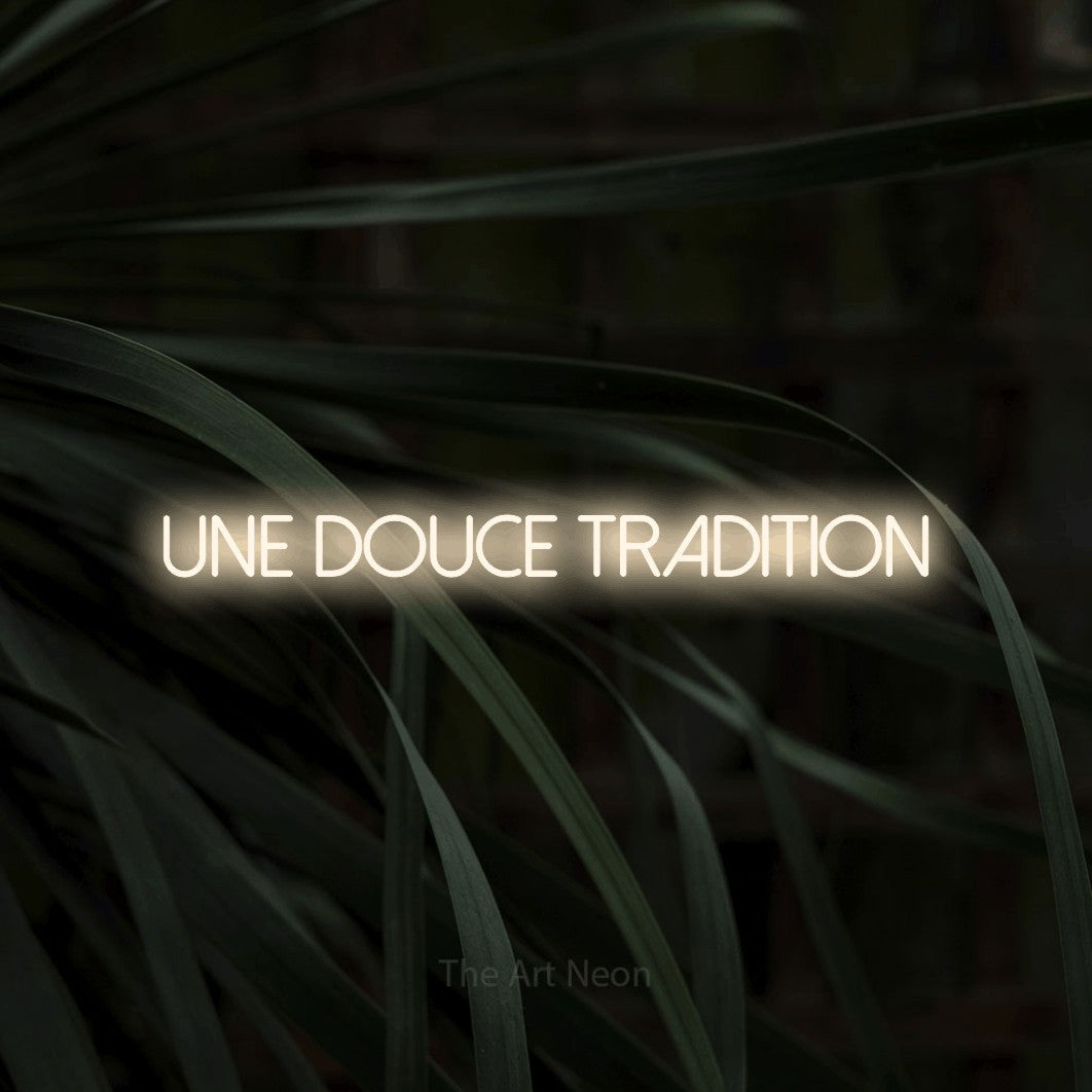 “Une douce tradition” LED neon sign in warm white, with thin sans-serif lettering, shown against a close-up of dark foliage.