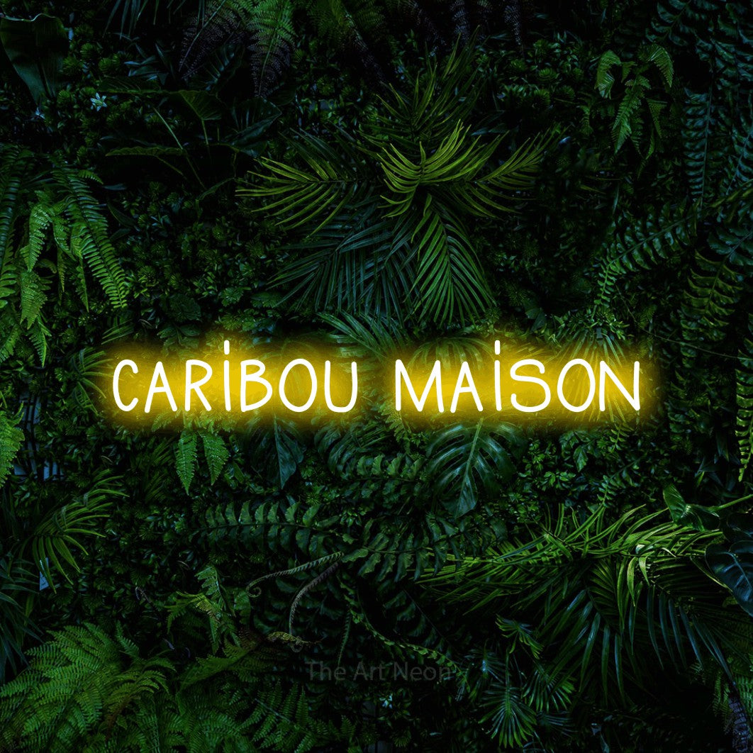 “Caribou maison” LED neon sign in yellow, with narrow hand-drawn lettering, shown against dense tropical foliage.