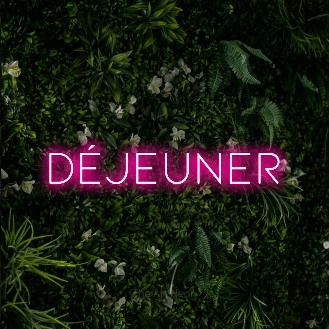 “Déjeuner” LED neon sign in hot pink, with thin, widely spaced lettering, shown against a dense green plant wall.