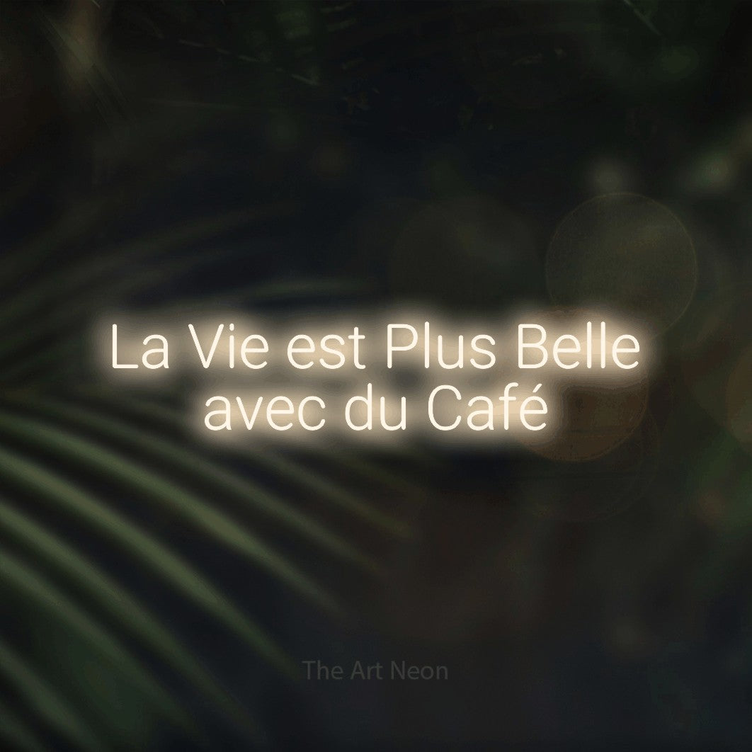“La vie est plus belle avec du café” LED neon sign in warm white, with rounded sans-serif lettering across 2 lines, shown against a blurred palm and bokeh-light backdrop.