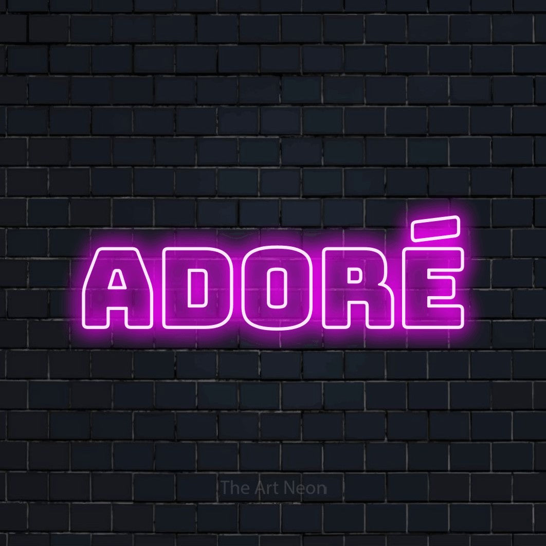 “Adoré” LED neon sign in pink, with bold outline lettering, shown against a dark navy brick wall.