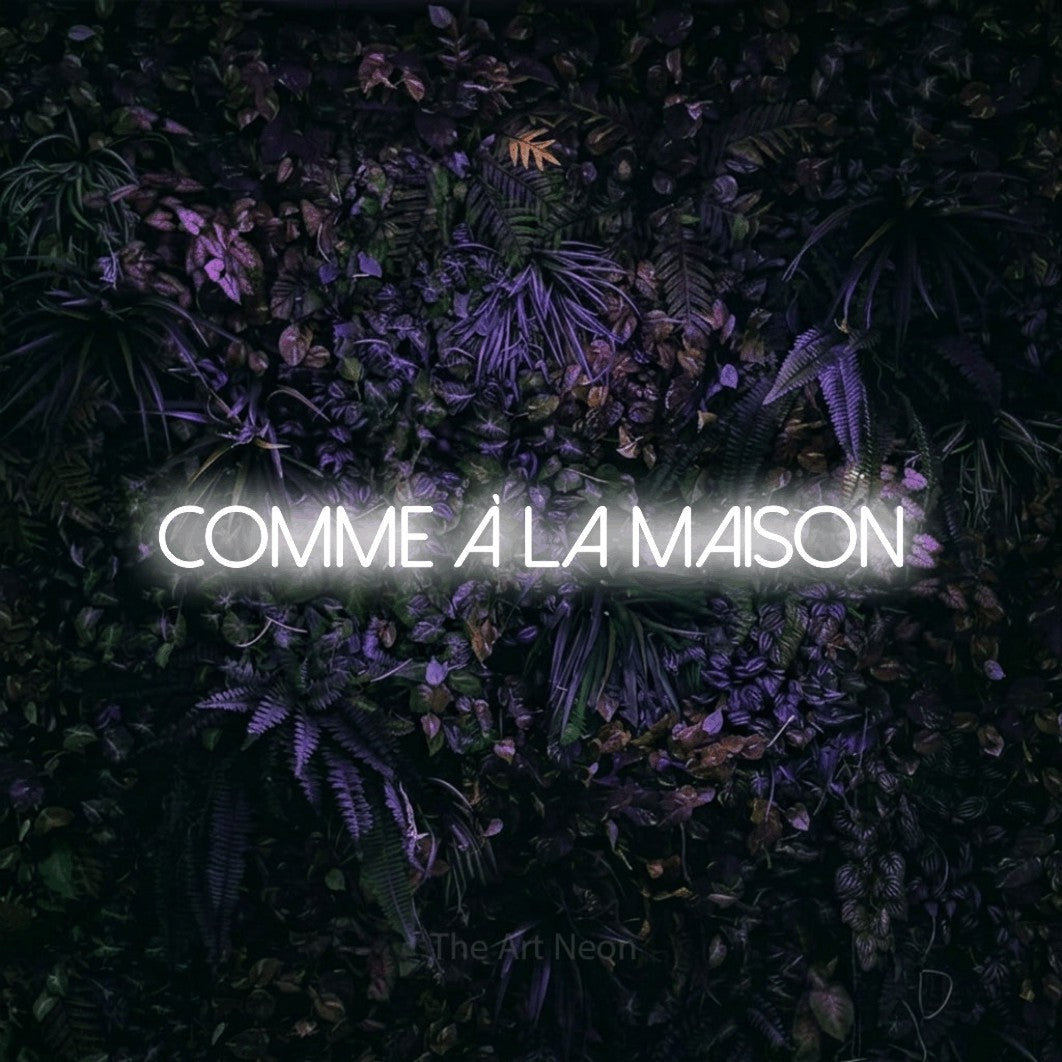 “Comme à la maison” LED neon sign in cool white, with thin sans-serif lettering, shown against a dark wall with purple flowers.