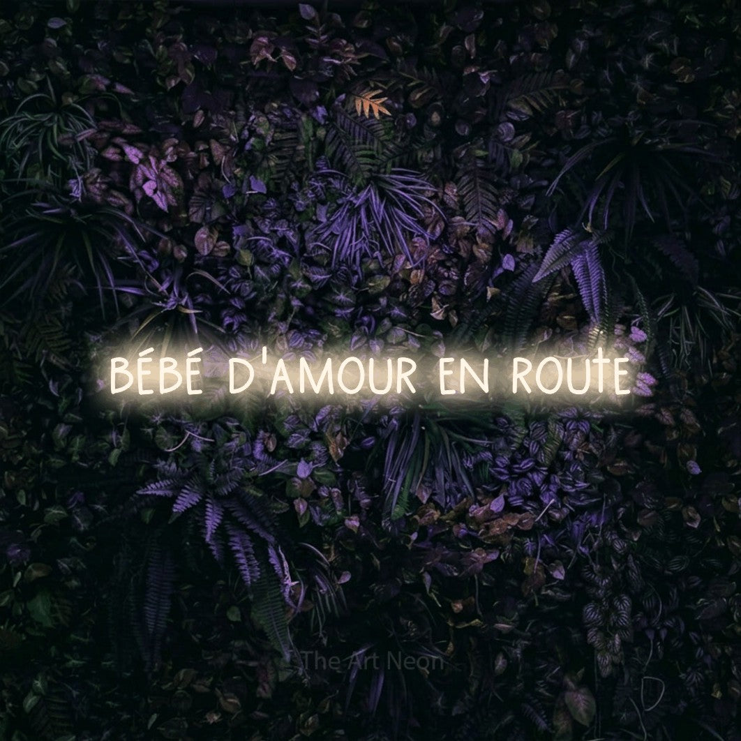 “Bébé d'amour en route” LED neon sign in warm white, with narrow hand-drawn lettering, shown against a dark wall with purple flowers.