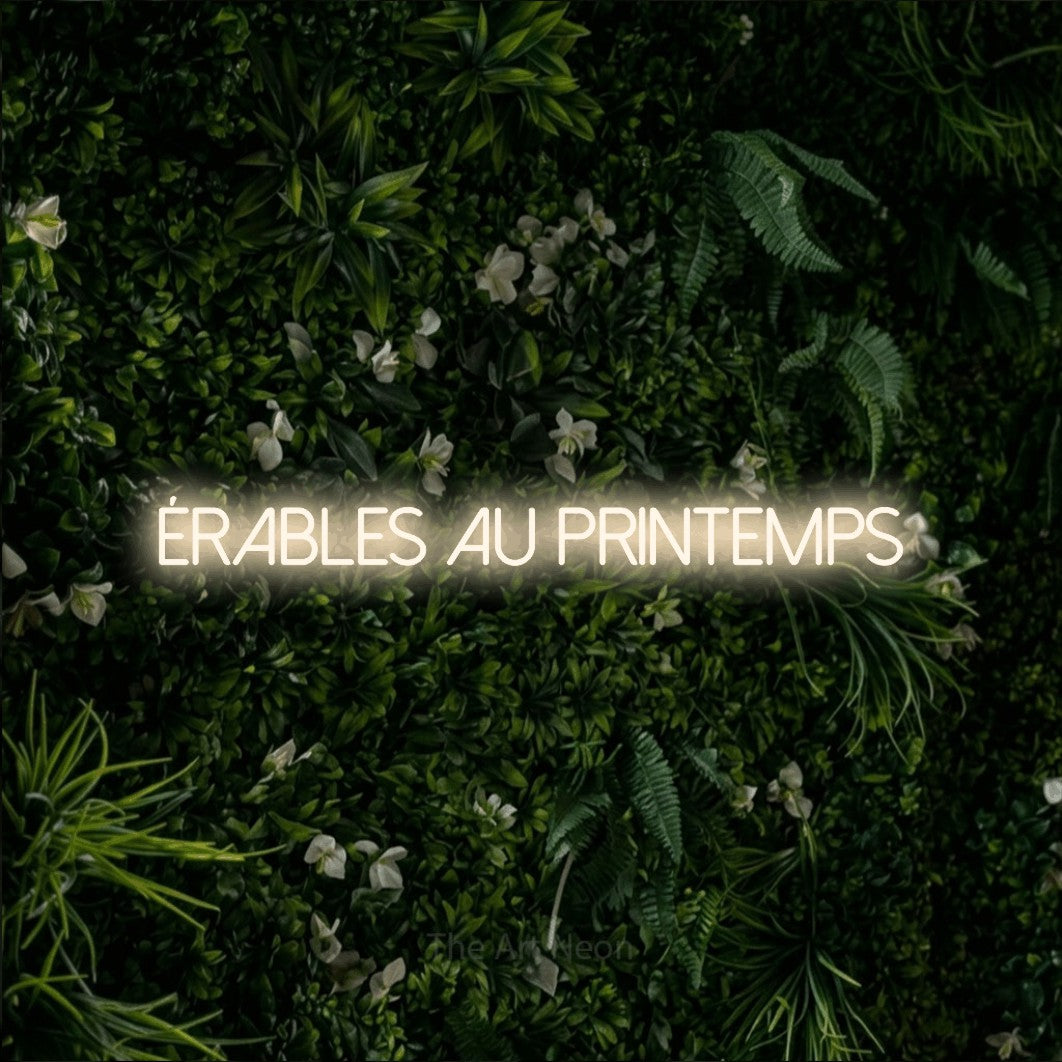 “Érables au printemps” LED neon sign in warm white, with thin sans-serif lettering, shown against a dense green plant wall.