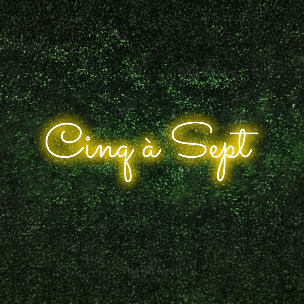 “Cinq à sept” LED neon sign in yellow, with looped cursive lettering, shown against a textured green hedge.