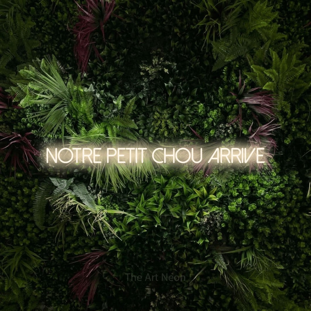 Décoration murale en néon LED « Notre petit chou arrive » en blanc chaud, en lettres fines sans empattement, installée sur un mur végétal aux fleurs roses, pour une fête prénatale.