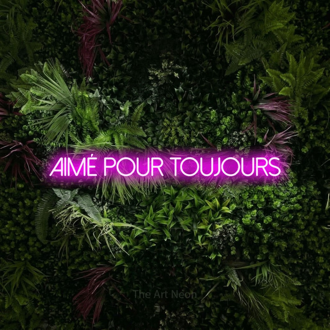 “Aimé pour toujours” LED neon sign in pink, with thin sans-serif lettering, shown against a plant wall with pink flowers.