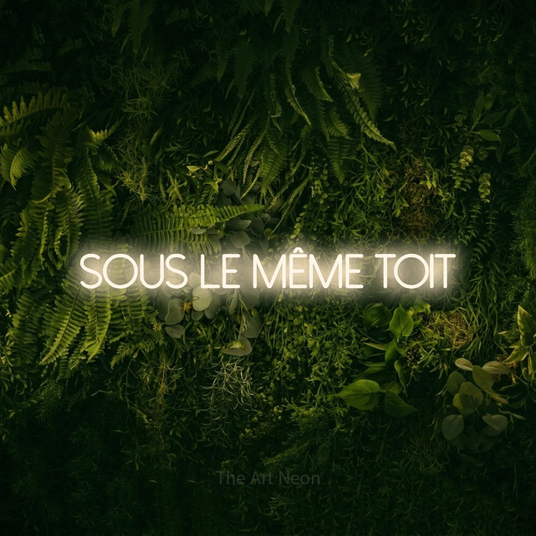 “Sous le même toit” LED neon sign in warm white, with thin sans-serif lettering, shown against a dark moss and forest backdrop.