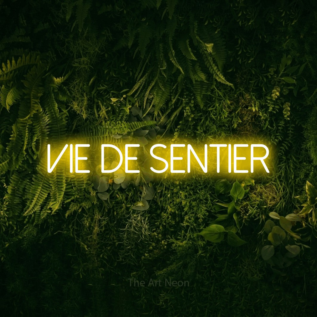 “Vie de sentier” LED neon sign in yellow, with thin sans-serif lettering, shown against a dark moss and forest backdrop.