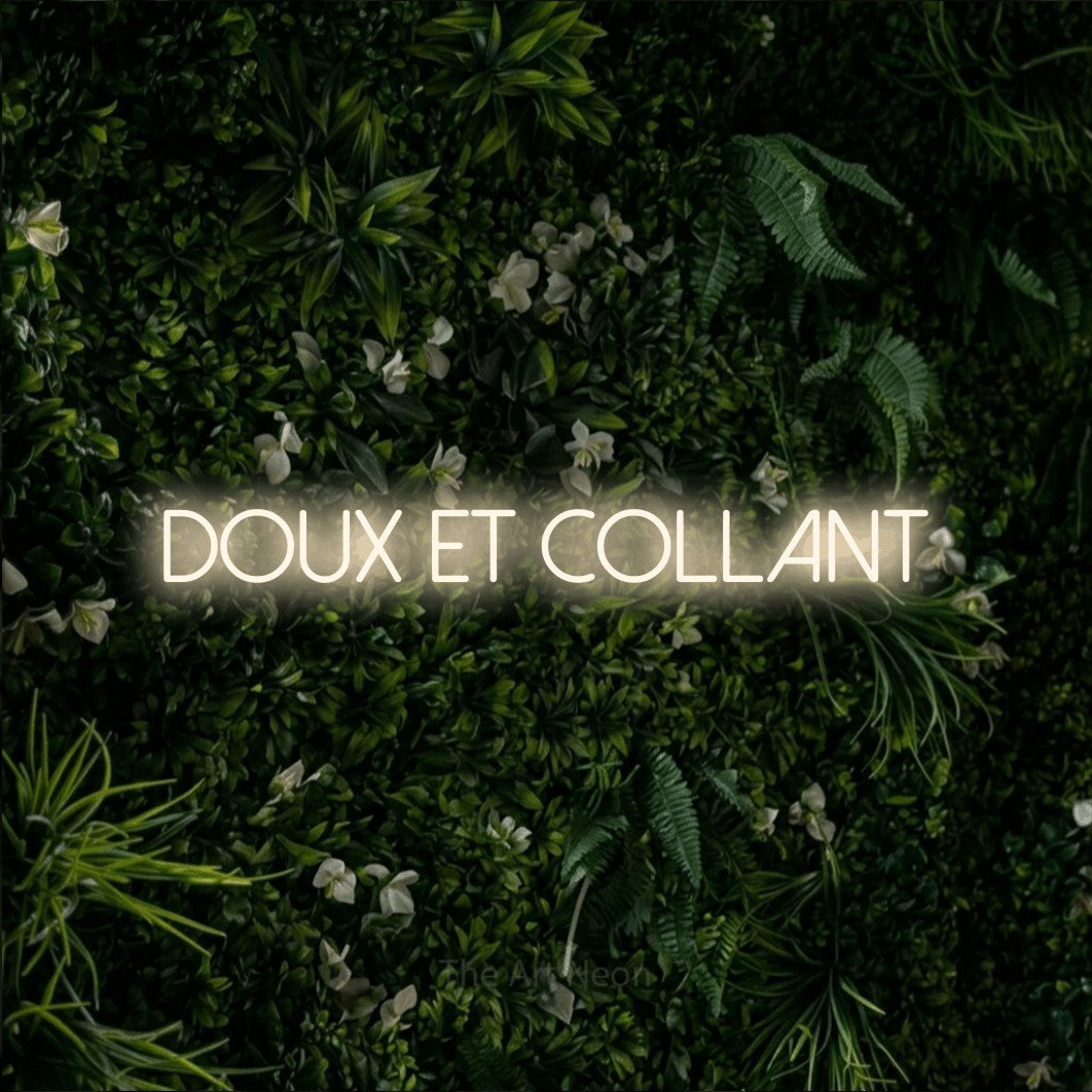 “Doux et collant” LED neon sign in warm white, with thin sans-serif lettering, shown against a dense green plant wall.