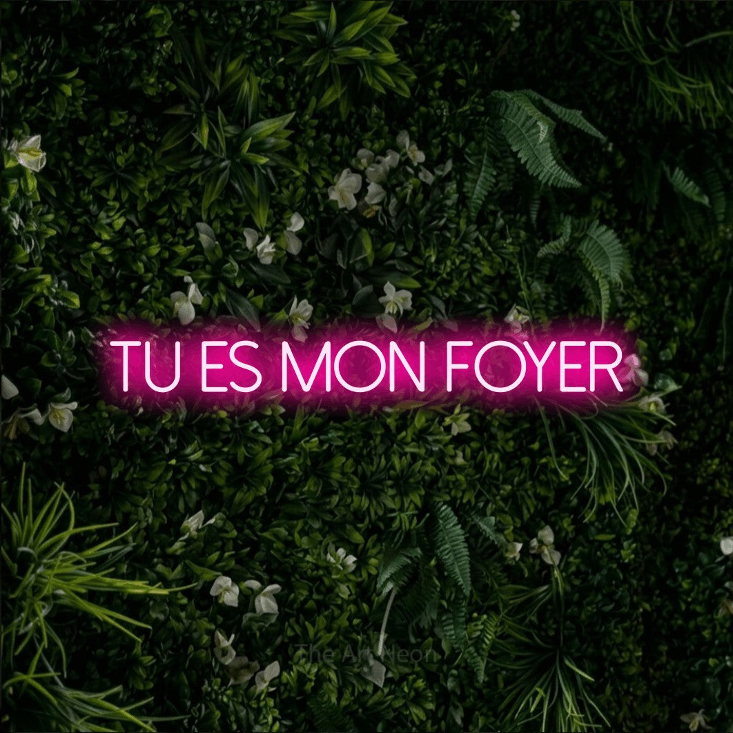 “Tu es mon foyer” LED neon sign in hot pink, with thin sans-serif lettering, shown against a dense green plant wall.