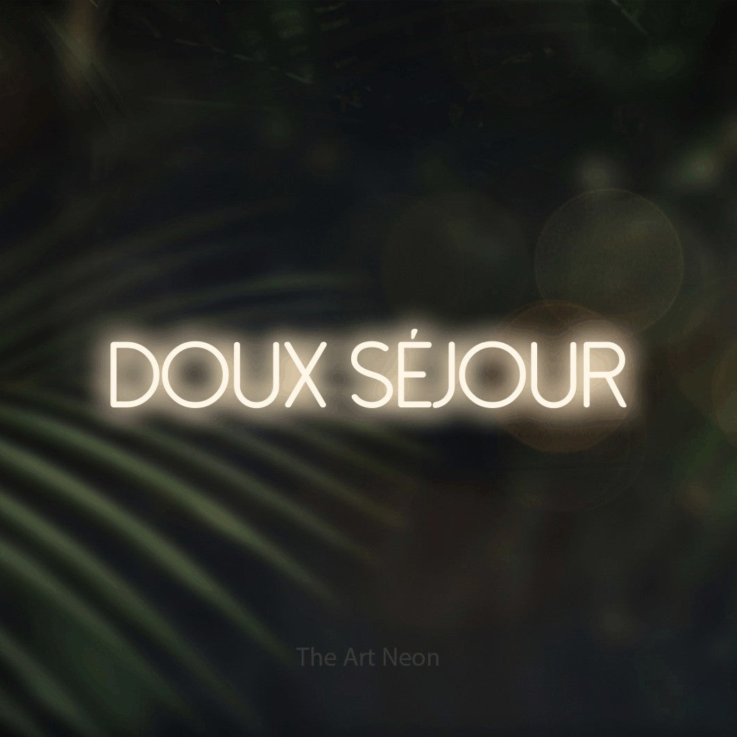 “Doux séjour” LED neon sign in warm white, with thin sans-serif lettering, shown against a blurred palm and bokeh-light backdrop.