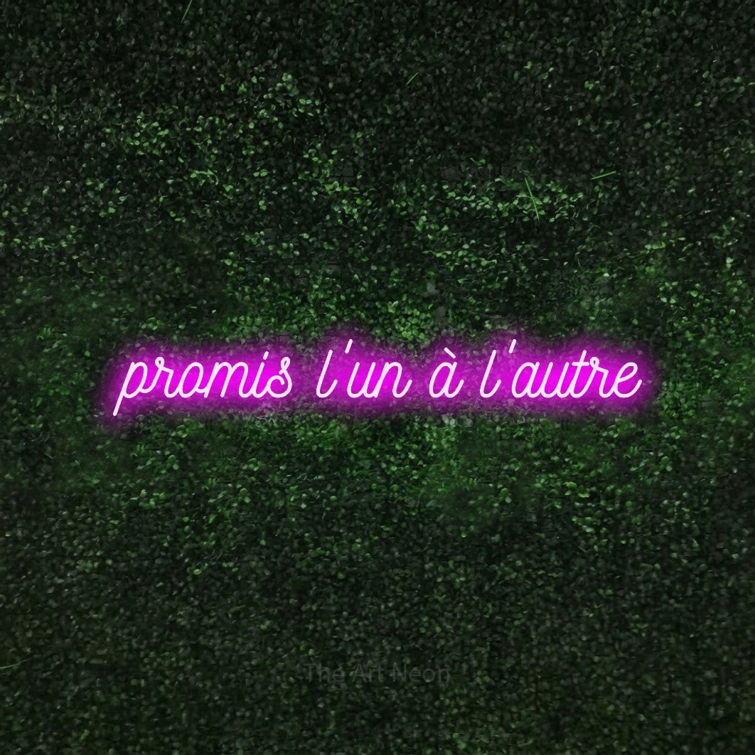 “Promis l'un à l'autre” LED neon sign in pink, with ornate cursive lettering, shown against a textured green hedge.