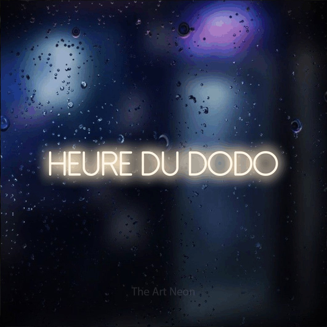 “Heure du dodo” LED neon sign in warm white, with thin sans-serif lettering, shown against glass with blue and purple reflections.