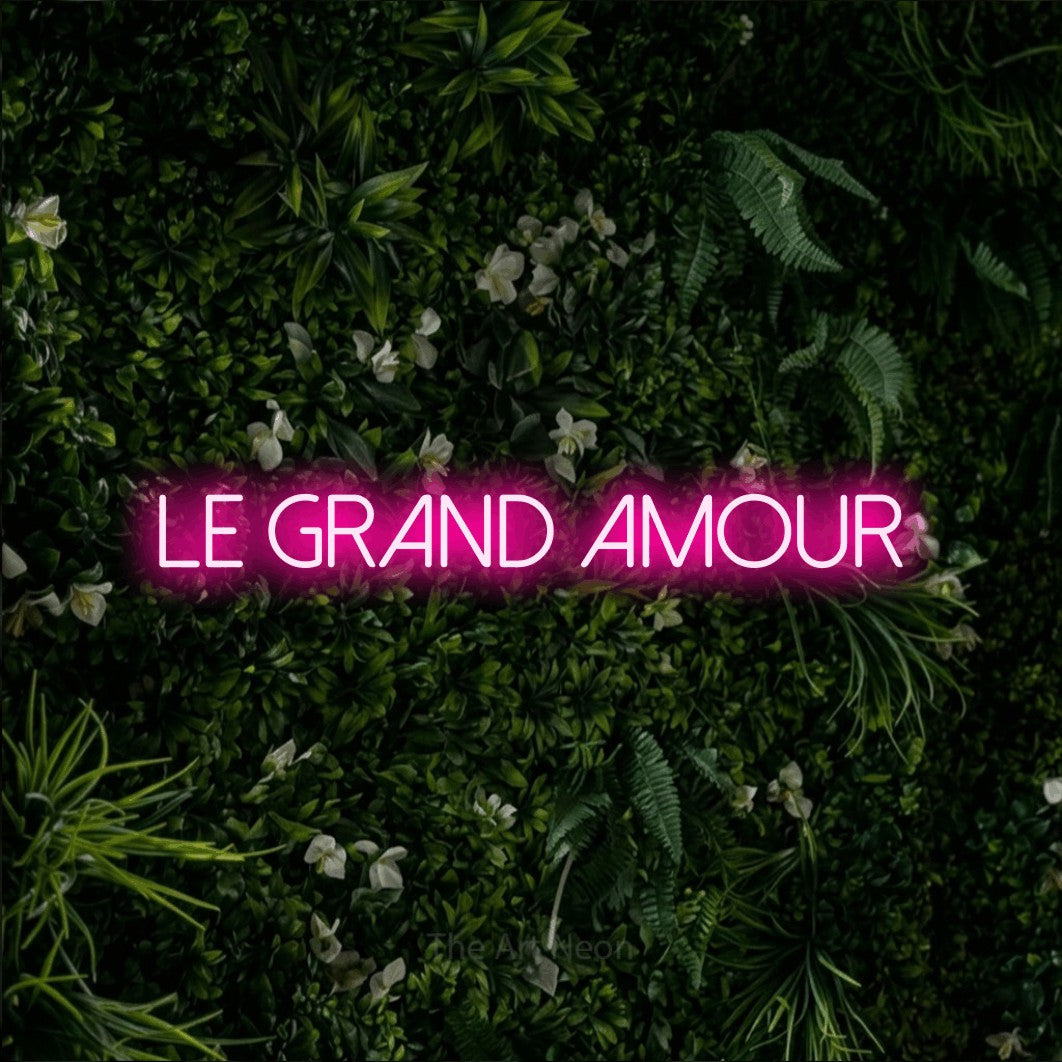“Le grand amour” LED neon sign in hot pink, with thin sans-serif lettering, shown against a dense green plant wall.