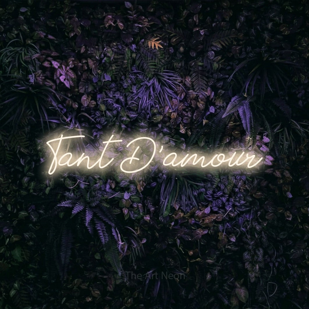 “Tant d'amour” LED neon sign in warm white, with cursive lettering, shown against a dark wall with purple flowers.