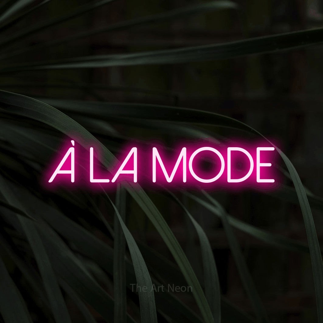 “À la mode” LED neon sign in hot pink, with thin sans-serif lettering, shown against a close-up of dark foliage.
