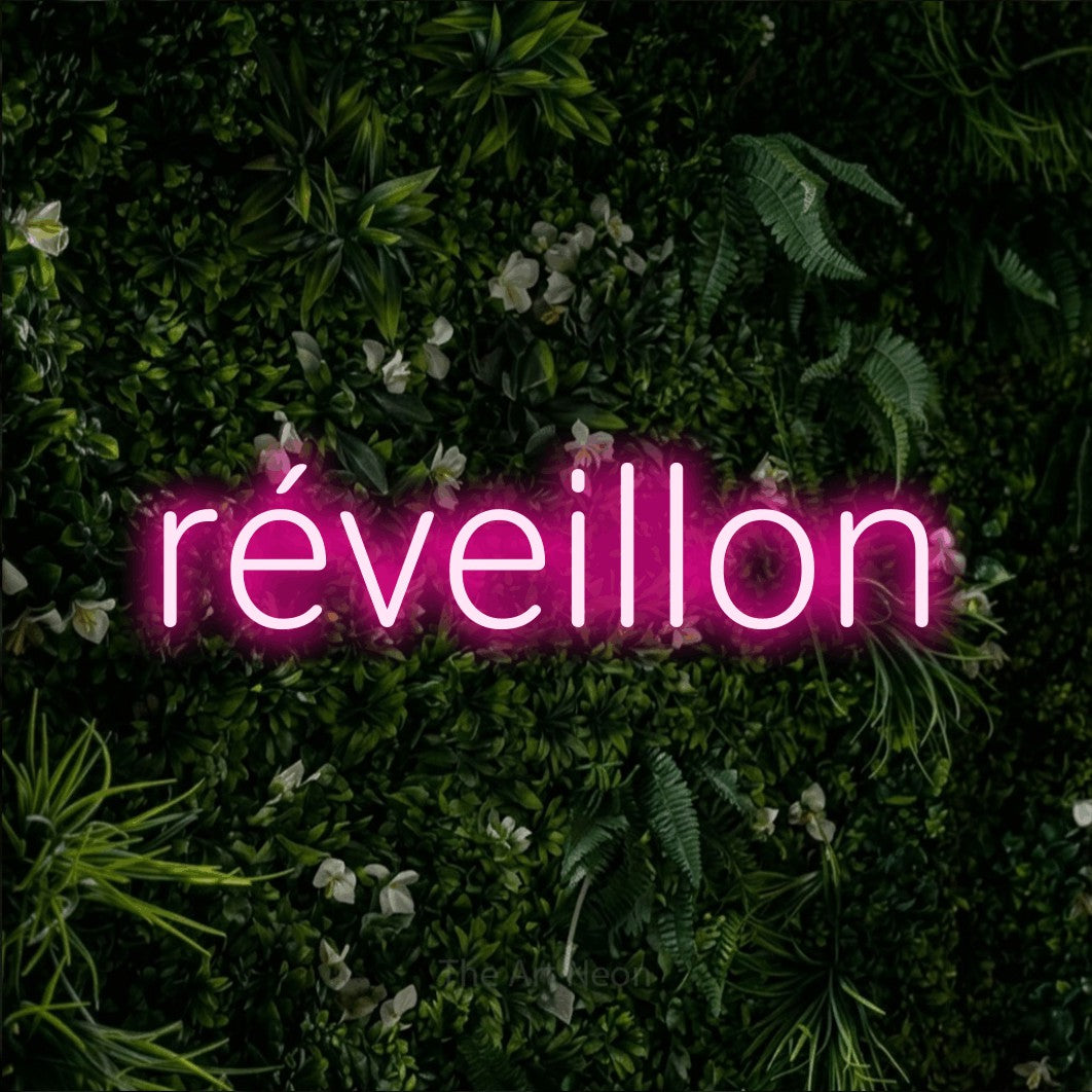 “Réveillon” LED neon sign in hot pink, with rounded sans-serif lettering, shown against a dense green plant wall.
