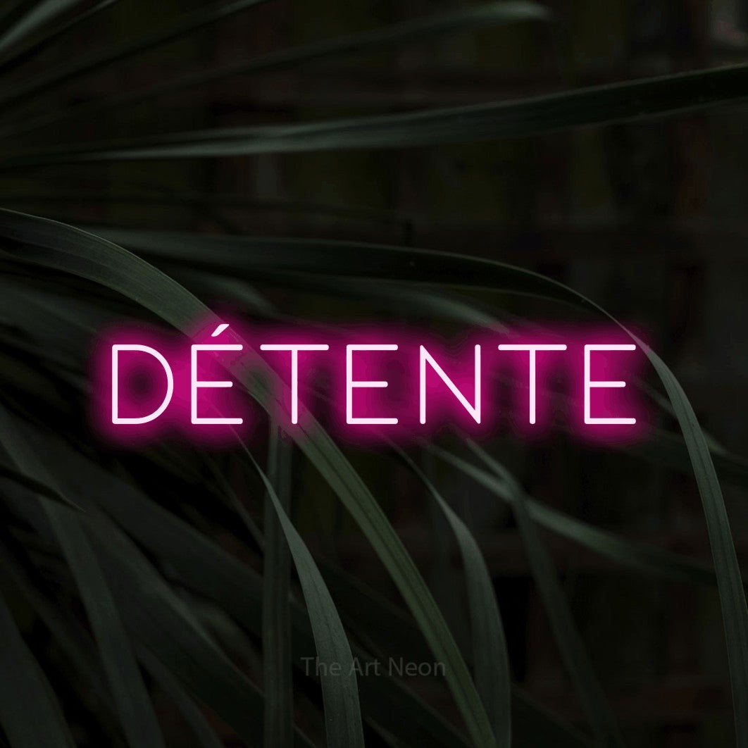 “Détente” LED neon sign in hot pink, with thin, widely spaced lettering, shown against a close-up of dark foliage.