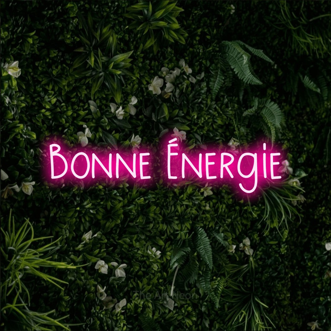Néon LED « Bonne énergie » en rose vif, en lettres étroites dessinées à la main, mis en scène sur un mur végétal vert et dense, pour un salon.