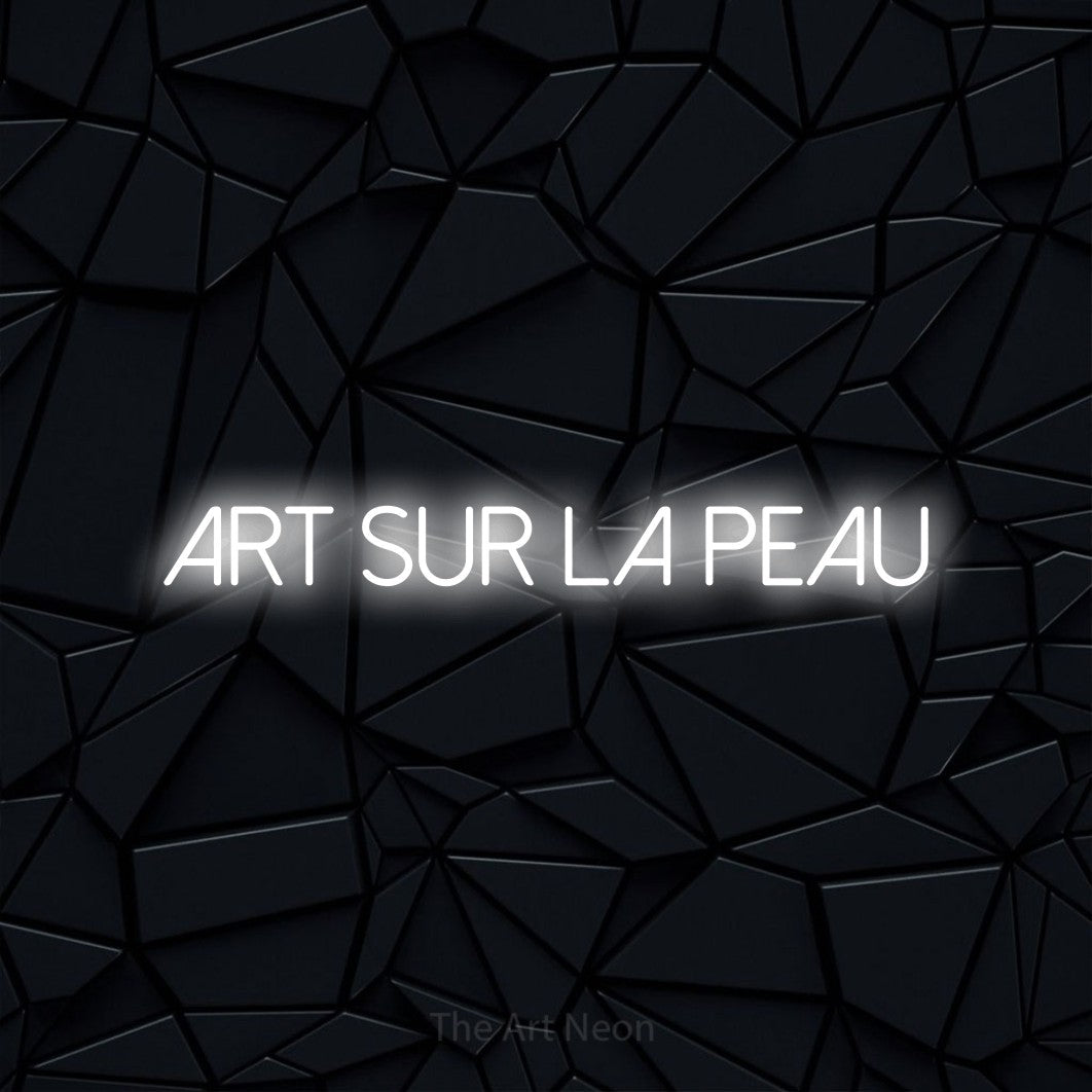 “Art sur la peau” LED neon sign in cool white, with thin sans-serif lettering, shown against a black polygonal surface.