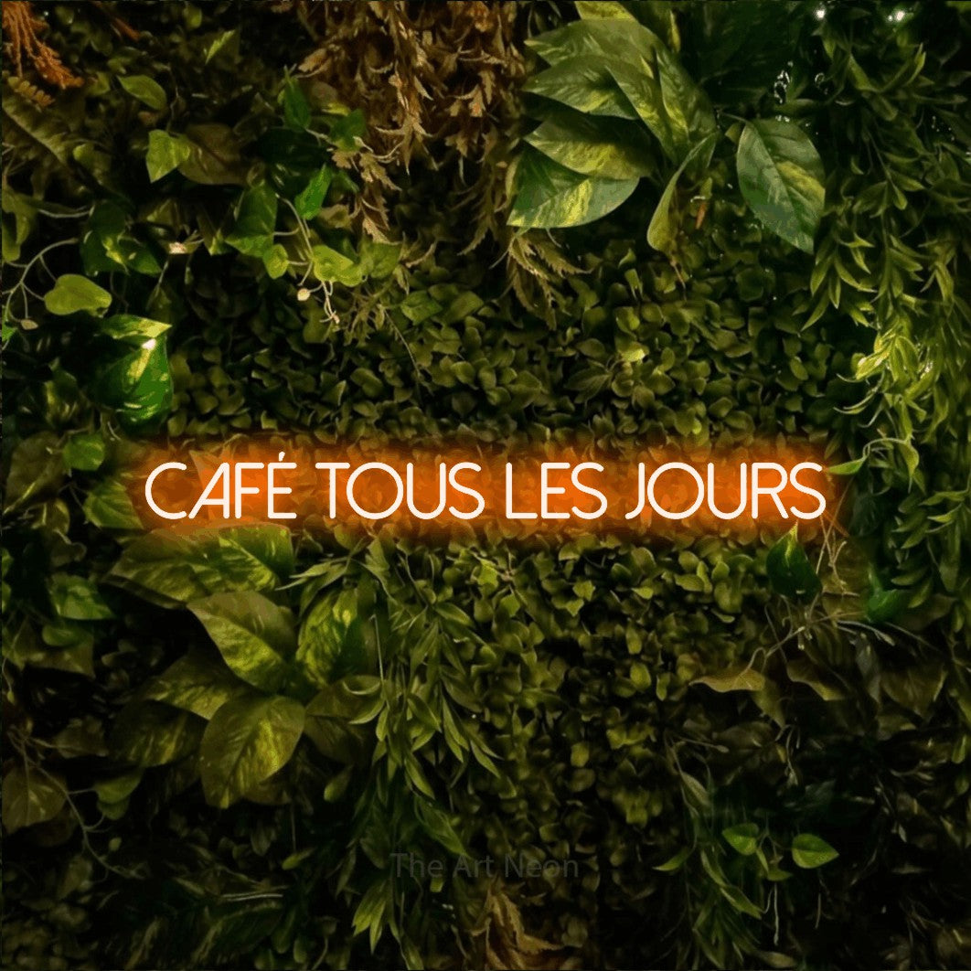 “Café tous les jours” LED neon sign in orange, with thin sans-serif lettering, shown against bright green and yellow foliage.
