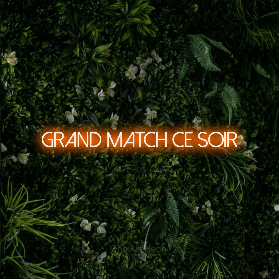 “Grand match ce soir” LED neon sign in orange, with thin sans-serif lettering, shown against a dense green plant wall.