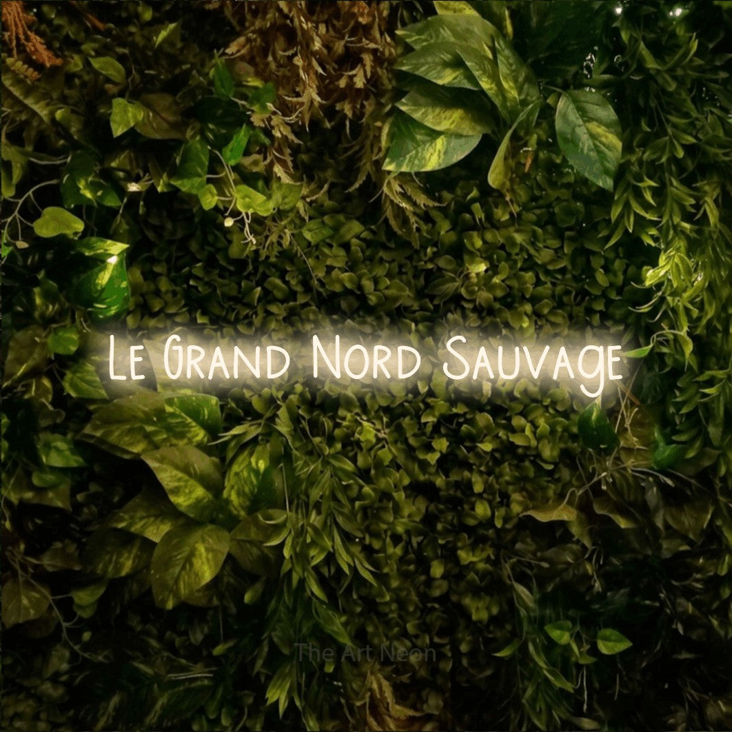 Lettrage en néon LED « Le grand nord sauvage » en blanc chaud, en lettres étroites dessinées à la main, présenté sur un feuillage vert et jaune vif, pour un décor de plein air canadien.