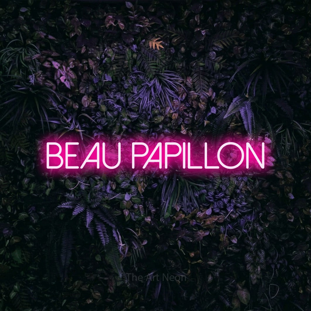 “Beau papillon” LED neon sign in hot pink, with thin sans-serif lettering, shown against a dark wall with purple flowers.