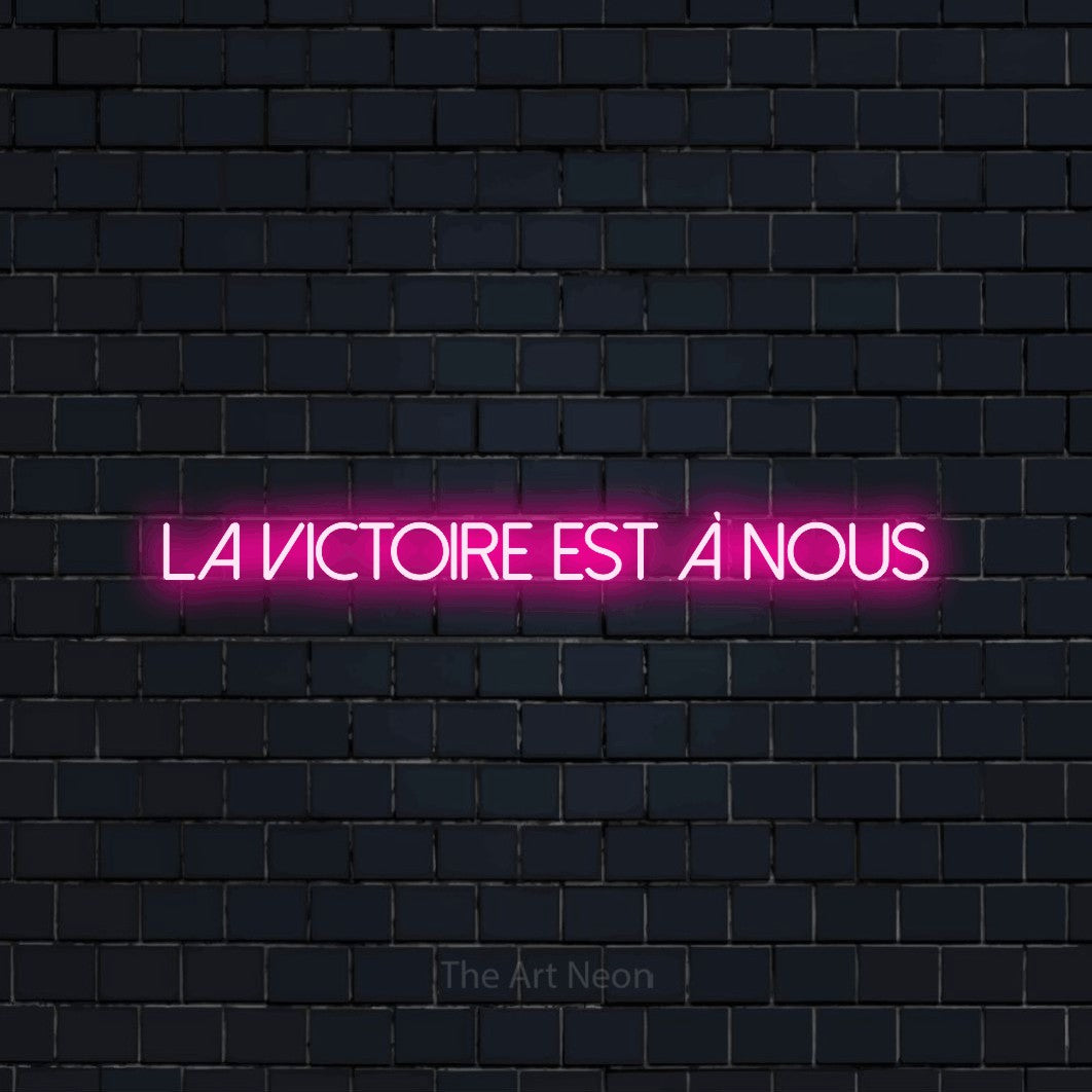 “La victoire est à nous” LED neon sign in hot pink, with thin sans-serif lettering, shown against a dark navy brick wall.