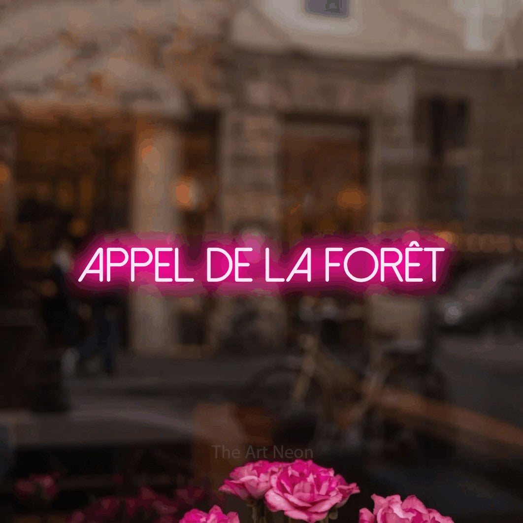 “Appel de la forêt” LED neon sign in hot pink, with thin sans-serif lettering, shown against a blurred café-bar interior.