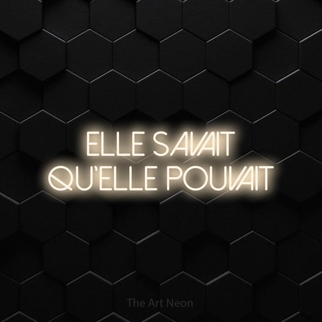 “Elle savait qu'elle pouvait” LED neon sign in warm white, with thin sans-serif lettering across 2 lines, shown against black hexagonal wall tiles.