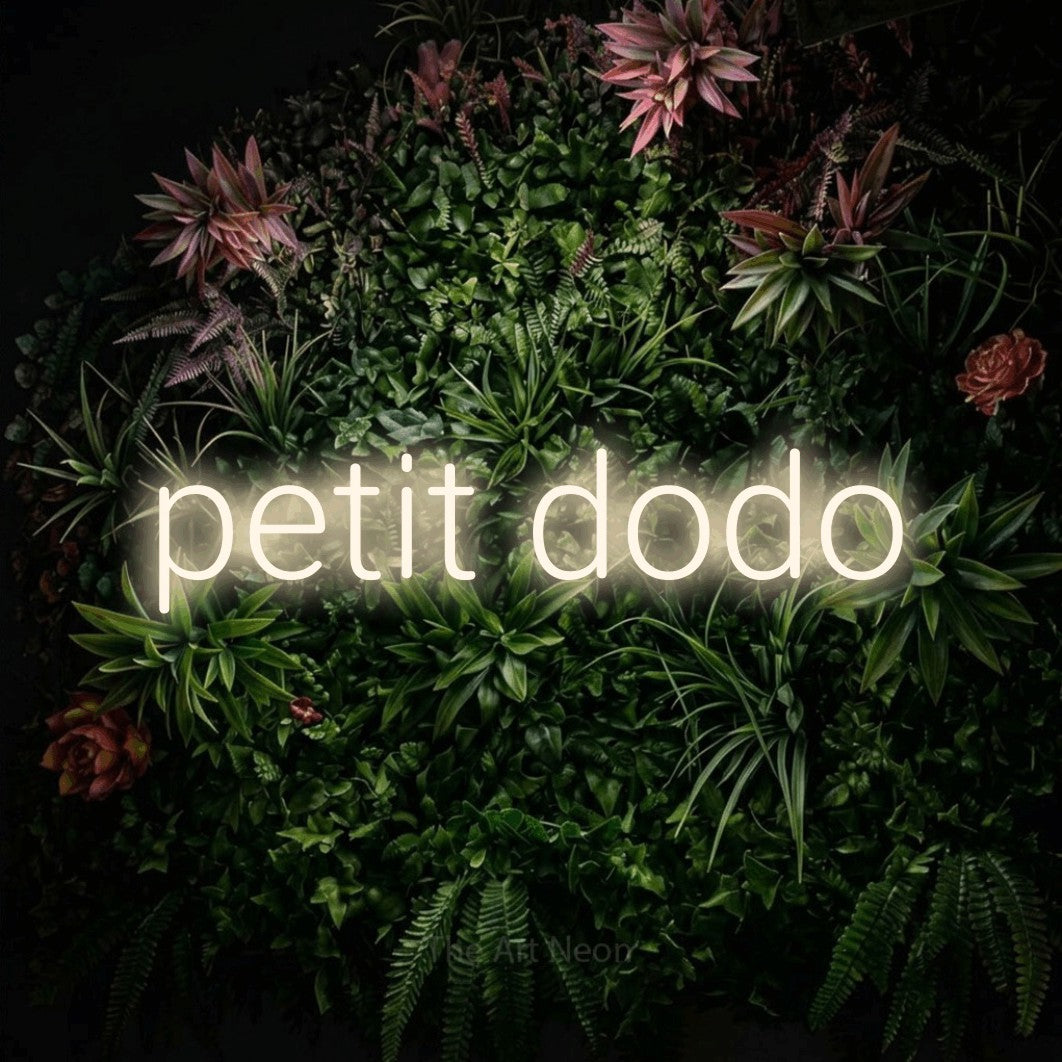“Petit dodo” LED neon sign in warm white, with rounded sans-serif lettering, shown against a flowering plant wall.