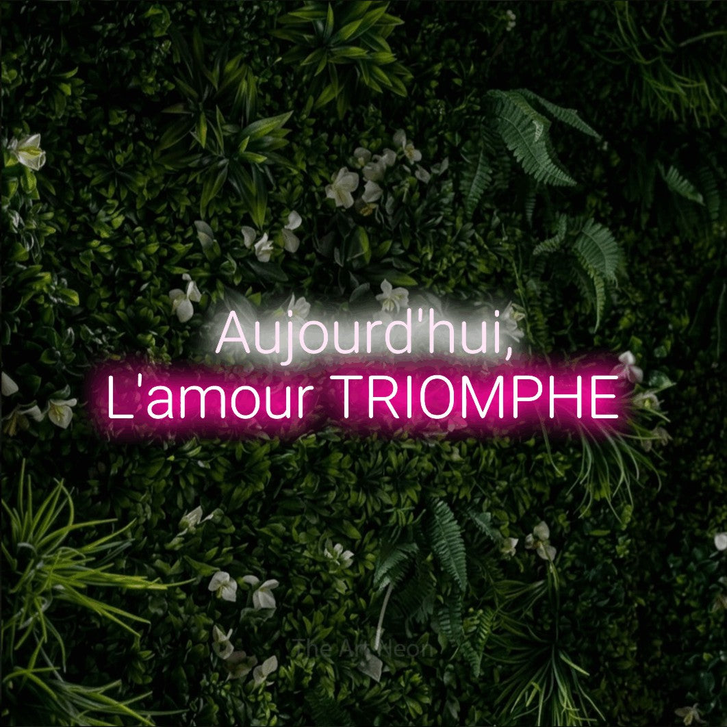“Aujourd'hui, l'amour triomphe” LED neon sign in cool white on the upper line and hot pink on the lower line, with rounded sans-serif lettering across 2 lines, shown against a dense green plant wall.